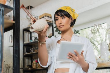 Young asian brunette artisan in workwear and headscarf holding digital tablet and looking at clay sculpture while standing in blurred ceramic workshop at background, process of pottery making clipart