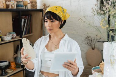 Young brunette asian artisan in headscarf and workwear holding clay sculpture and using digital tablet while standing in ceramic workshop, creative process of pottery making clipart