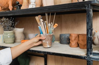 Cropped view of young craftswoman taking pottery tools from shelf with clay products and blurred lavender flowers at background in ceramic workshop, creative process of pottery making clipart