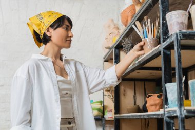 Atölye ve başörtüsü içinde genç Asyalı kadın sanatçı kil ürünlerinin yanındaki raflara çömlek aletleri koyuyor ve seramik atölyede çalışıyor, çömlek yapımında yaratıcı bir süreç.