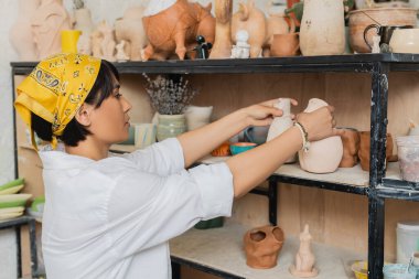 Side view of young asian female potter in headscarf and workwear putting clay product on shelf on rack while working in ceramic workshop, pottery studio scene with skilled artisan clipart