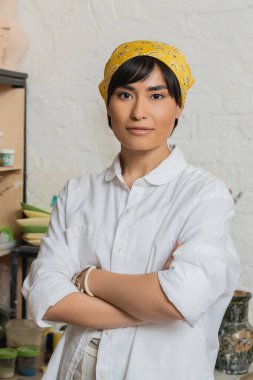 Genç Asyalı kadın çömlekçinin portresi başörtüsü ve atkı takmış kollarını kavuşturup seramik atölyede dururken kameraya bakıyor, yetenekli bir zanaatkarla çömlek stüdyosu sahnesi