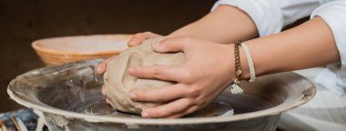 Cropped view of young female potter putting clay on pottery wheel while working near bowl in blurred ceramic workshop, artisan crafting ceramics in studio, banner  clipart