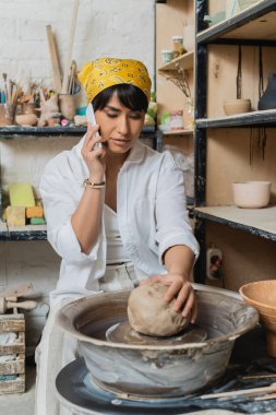 Young asian female artisan in headscarf and workwear talking on smartphone and putting clay on pottery wheel in ceramic workshop, artisan crafting ceramics in studio clipart