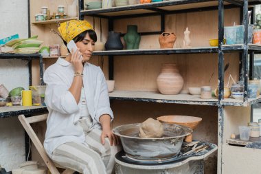 Young asian female potter in workwear and headscarf talking on smartphone while sitting near clay on pottery wheel and tools and working in pottery studio, artisan crafting ceramics in studio clipart