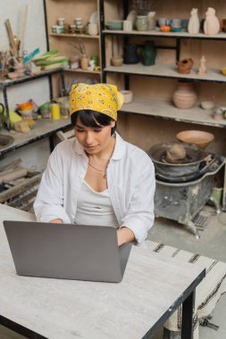 Masada otururken ve bulanık seramik atölyesinde çalışırken başörtüsü ve atkı kullanan Asyalı genç kadın zanaatkârın yüksek açılı görüntüsü, çömlek sanatçısı sanatını sergiliyor.