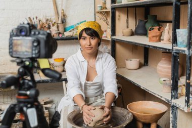 Genç esmer Asyalı esmer kadın başörtüsü ve iş kıyafetleri içinde çömlek çarkının üzerindeki kil pervazda bulanık dijital kamera yanında tripod ve rafta heykeller stüdyoda, çömlekçi sanatçısı sanatını sergiliyor.