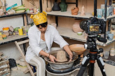 Young asian female artisan in workwear and headscarf molding clay on pottery wheel near blurred digital camera on tripod in ceramic workshop, pottery artist showcasing craft clipart