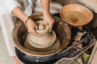 Top view of young female artisan molding wet clay on pottery wheel near tools and bowl with water in ceramic workshop, pottery studio workspace and craft concept clipart