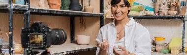 Cheerful young asian female potter talking to blurred digital camera while sitting near clay sculptures on rack in ceramic workshop, clay sculpting process concept, banner clipart