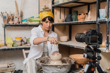 Young asian female potter in headscarf pouring water from sponge on clay on pottery wheel near blurred digital camera in ceramic workshop, clay sculpting process concept clipart