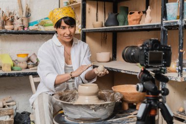 Positive asian female artist in headscarf holding wet clay and looking at digital camera on tripod near pottery wheel in art workshop, clay sculpting process concept clipart