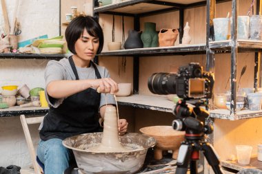 Young brunette asian female artisan in apron pouring water from sponge on wet clay and pottery wheel near digital camera on tripod in studio at background, pottery tools and equipment clipart