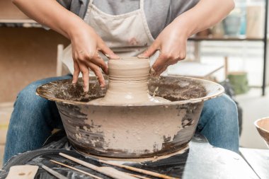 Cropped view of blurred female artisan in apron shaping wet clay on pottery wheel near tools on table in ceramic art workshop ay background, clay sculpting process concept clipart