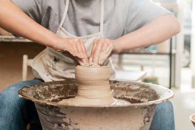 Önlüklü genç bayan çömlekçinin ıslak çamur kalıbını çıkarması ve arka planda bulanık sanat seramik stüdyosunda çömlek çarkı ile çalışması, kil heykel süreci konsepti.