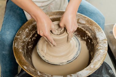 High angle view of young female potter in apron shaping wet clay while working on spinning pottery wheel in art workshop at background, skilled pottery making concept clipart