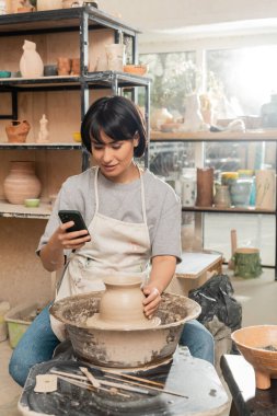 Smiling asian brunette craftswoman in apron using smartphone while working with clay on pottery wheel near wooden tools and bowl in ceramic workshop, pottery creation process clipart