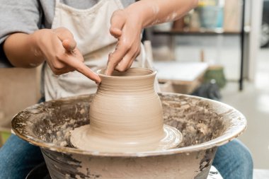 Önlüklü genç kadın zanaatkarın ıslak çamurdan vazo yapmasını ve bulanık seramik atölyesinde dönen çömlek çarkı ile çalışmasını, çömlek oluşturma işlemini.