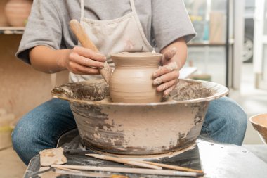 Cropped view of young female artisan in apron making shape of clay vase with wooden tool on spinning pottery wheel in blurred ceramic workshop at background, pottery creation process clipart