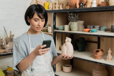 Önlüklü Asyalı genç esmer kadın esmer. Akıllı telefon kullanıyor ve seramik heykel tutuyor. Arka planda seramik atölyesinde seramik ürünlerin olduğu rafın yanında duruyor.