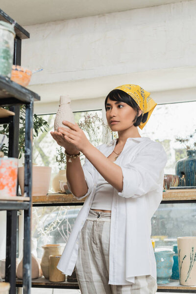 Brunette asian female artist in headscarf and workwear holding clay product while standing near shelves in blurred pottery studio, pottery studio with artisan at work