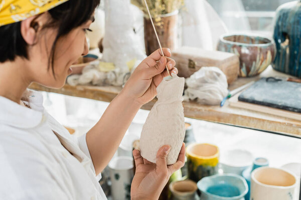 Side view of smiling blurred young asian female artist holding wooden stick and clay product while working in blurred pottery class at background, pottery studio with artisan at work