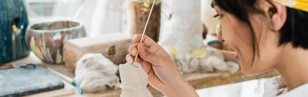 Side view of young smiling blurred asian artist holding wooden stick and clay sculpture while working in blurred pottery class at background, pottery studio with artisan at work, banner 