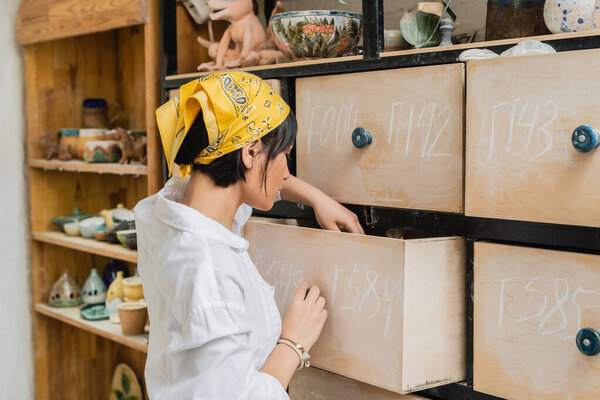 Side view of young asian female artist in headscarf and workwear opening cupboard while working in blurred pottery workshop, pottery workshop with skilled artisan