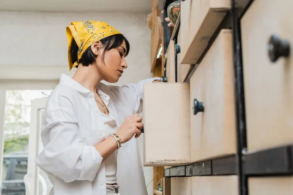 stock image Side view of young brunette asian female artist in workwear and headscarf opening cupboard while working in blurred pottery class, pottery workshop with skilled artisan