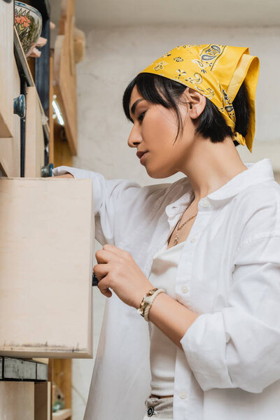 Side view of young brunette asian artist in headscarf and workwear opening cupboard while working in pottery studio, pottery workshop with skilled artisan