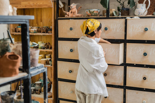 Side view of young brunette asian craftswoman in headscarf and workwear opening cupboard while standing and working in ceramic studio, pottery workshop with skilled artisan