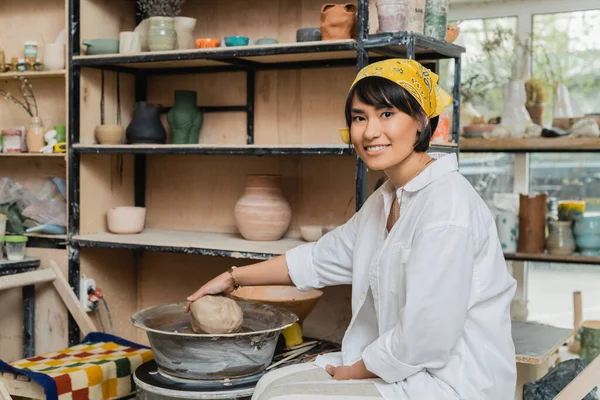 頭皮のスカーフや作業着を持つ陽気なブルネットのアジアの女性の陶芸家粘土を保持し セラミックワークショップで陶器ホイールの近くのカメラを見て 陶器作りの職人技 — ストック写真
