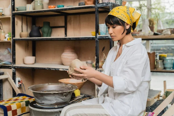ヘッドスカーフの若いアジアの女性職人や粘土を保持しながら 陶芸工房で陶芸ホイールの近くで作業している作業着 陶器作りの職人技 — ストック写真