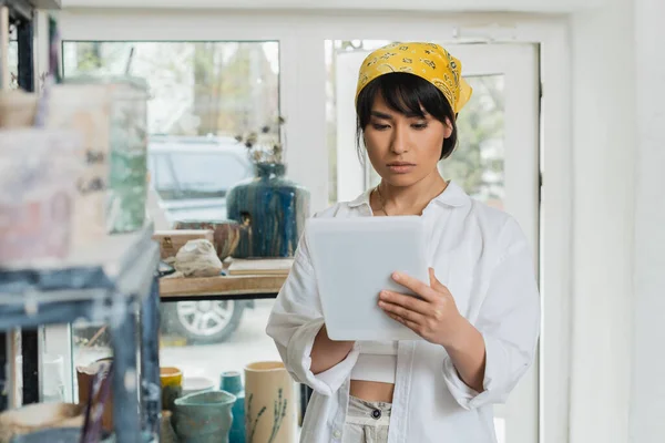 stock image Portrait of young brunette asian female artisan in headscarf and workwear using digital tablet while standing in blurred ceramic workshop at background, creative process of pottery making