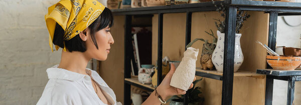 Side view of young brunette asian female potter in headscarf and workwear holding clay sculpture near shelves in ceramic workshop at background, creative process of pottery making, banner 