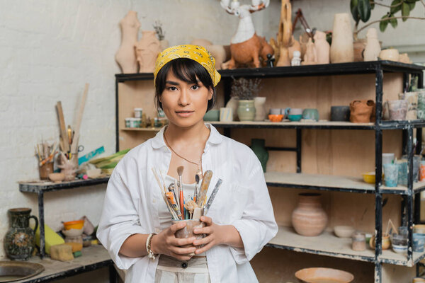 Young brunette asian female artisan in workwear and headscarf holding pottery tools and looking at camera while standing in ceramic workshop, creative process of pottery making