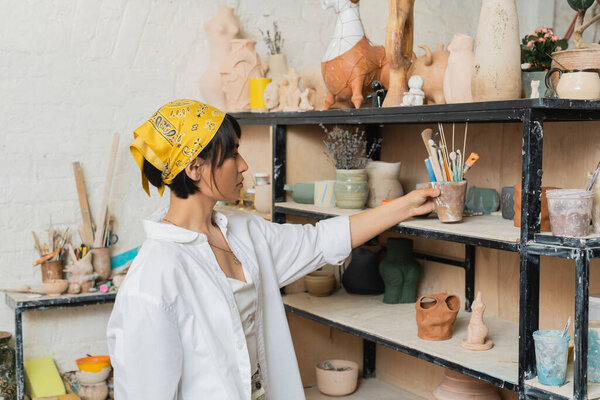 Side view of young asian artisan in headscarf and workwear taking pottery tools from shelf with clay products in ceramic studio, creative process of pottery making