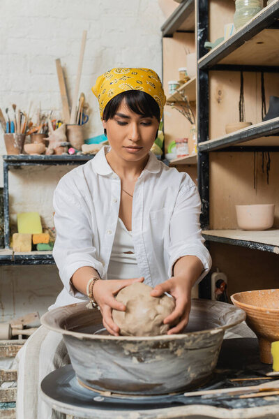 Young brunette asian artisan in headscarf and workwear holding clay while working with pottery wheel near rack and blurred pottery tools in workshop, pottery studio scene with skilled artisan