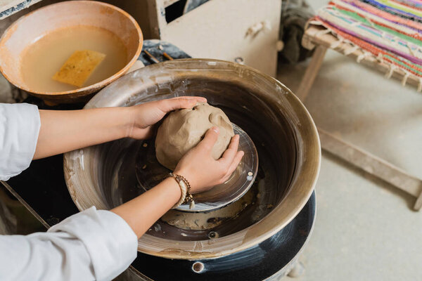 High angle view of young female artisan in workwear molding clay while working with pottery wheel near bowl with water in workshop, pottery studio scene with skilled artisan