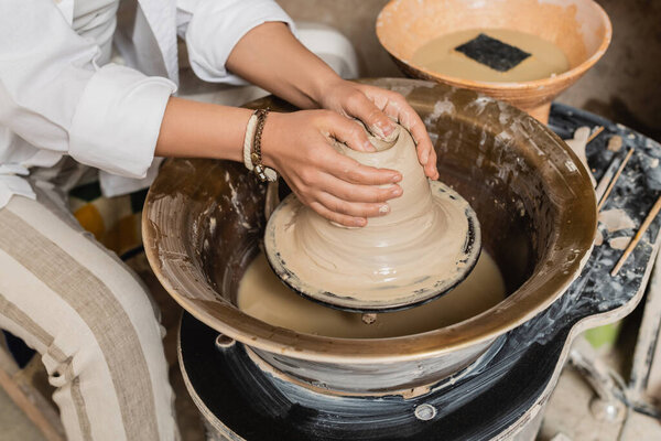 Cropped view of young artisan in workwear shaping wet clay while working on pottery wheel near blurred bowl with water and sponge at background, pottery studio workspace and craft concept