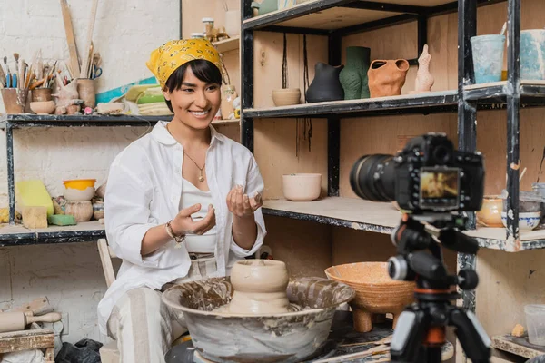 stock image Positive young asian artisan in headscarf and workwear looking at blurred digital camera on tripod near clay on pottery wheel and rack with sculptures in workshop, clay sculpting process concept