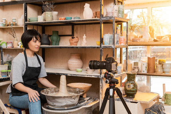 Young asian brunette female potter in apron looking at digital camera on tripod while sitting near pottery wheel and sculptures on rack in ceramic workshop, pottery tools and equipment