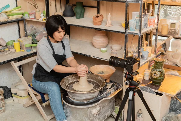 stock image Young asian female artisan in apron shaping wet clay on pottery wheel near bowl with water and tools near digital camera in ceramic workshop at background, pottery tools and equipment