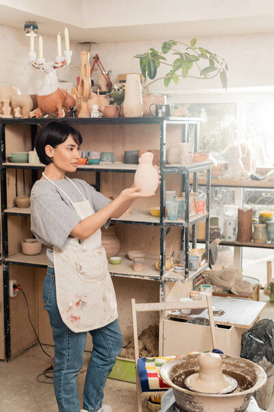 Young brunette asian female artist in apron holding sculpture while standing near clay on pottery wheel and rack in blurred ceramic studio at sunset, clay sculpting process concept