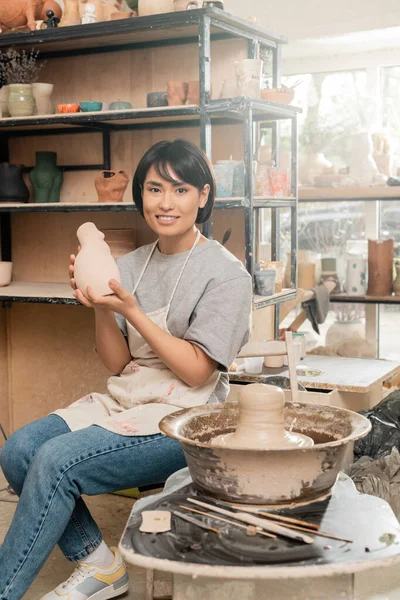 陶彫刻を持つエプロン姿の若いアジアの女性職人を笑顔にし ぼやけた工房の陶器の車輪の上にぬれた粘土の近くに座りながらカメラを見る 粘土彫刻のプロセスコンセプト — ストック写真