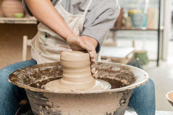 Cropped view of blurred female ceramicist in apron molding and shaping wet clay while working with spinning pottery wheel in art workshop, skilled pottery making concept