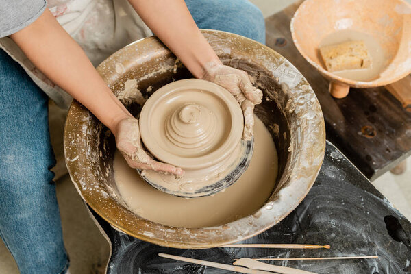 Top view of young craftswoman in apron making shape of wet clay on pottery wheel near bowl with sponge and tools on table in art studio, skilled pottery making concept