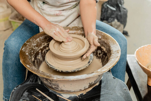 Cropped view of young female artist in apron molding clay and making shape while working on pottery wheel near wooden tools in art studio, skilled pottery making concept