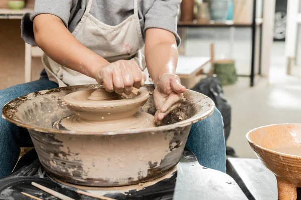 陶磁器工房の水で茶碗の近くの回転陶器の車輪の上に粘土の近くに湿ったスポンジを保持エプロンの若い女性の陶芸家の作物ビュー 陶器の作成プロセス — ストック写真