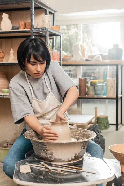 stock image Young brunette asian female artisan in apron shaping clay vase while working on pottery wheel near wooden tools and bowl in blurred ceramic workshop, pottery creation process